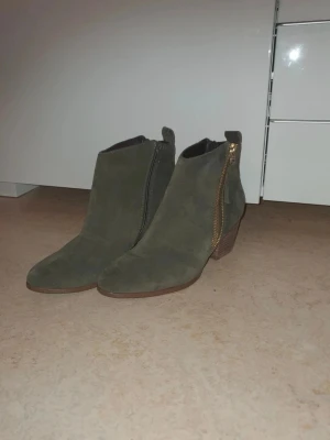 Olivgröna mockastövletter med klack - Snygga olivgröna stövletter i mocka med guldfärgade dragkedjor på sidan och låg träklack. Skorna har spetsig tå och stilren design som passar till många outfits. Perfekta för dig som vill ha en trendig och bekväm sko.