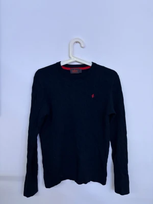 Morris stickad tröja - Snygg mörkblå kabelstickad tröja från Morriś med rund halsringning och röd broderad logga på bröstet. Tröjan har långa ärmar och klassisk passform, perfekt för dig som gillar stilrena och tidlösa plagg.