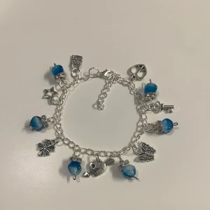 Silverfärgat armband med berlocker  - HANDGJORT armband i silverfärg med flera olika berlocker. Armbandet har blå pärlor som ger en färgklick och kedjan är justerbar. 