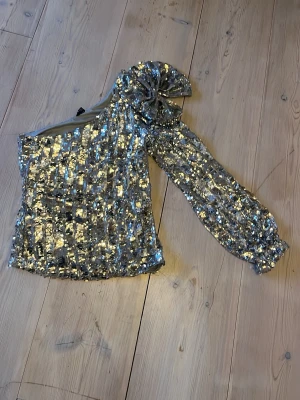 Glittrig topp - Säljer en glittrig silverfärgad topp med paljetter. Toppen har en oneshoulder-design med en lång puffärm och stor rosett på axeln. Från Vero Moda st xs använd 1 gång