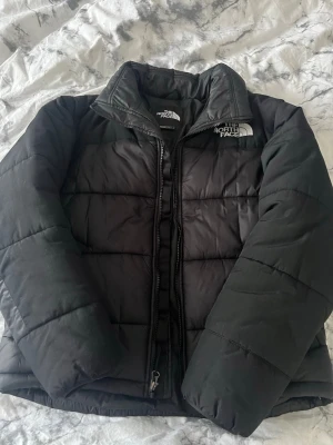 Svart pufferjacka från The North Face - Säljer en svart pufferjacka från The North Face med hög krage och dragkedja framtill. Jackan har ett quiltat mönster och logga på bröstet och ryggen. Perfekt för kalla dagar och riktigt snygg till streetwear-stilen. Jag säljer den eftersom den är för liten på mig.