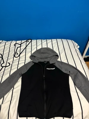 Svart och grå hoodie från FULLYPAID - Snygg hoodie med dragkedja från FULLYPAID. Tröjan är svart med grå ärmar och huva, samt har vit logga på bröstet. Perfekt för en chill och sportig stil. Mjukt material och två fickor framtill. Säljs ej längre av märket