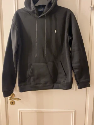 Svart Ralph Lauren hodie - Snygg svart hoodie från Polo Ralph Lauren med klassisk huva och dragsnören. Har en liten vit logga broderad på bröstet och en stor känguruficka framtill. Tillverkad i mjukt material som känns skönt mot huden. Perfekt för en avslappnad och stilren look. Storlek XL men passar L
