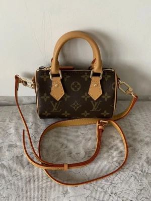 Louis Vuitton Nano speedy - Snygg handväska från Louis Vuitton i klassiskt monogrammönster. Väskan är brun med ljusbruna läderdetaljer och guldfärgade metalldelar. Den har rund form, dubbla handtag och dragkedja upptill. Perfekt för dig som gillar lyxiga accessoarer med ikonisk design.
