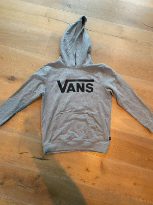 Grå hoodie från Vans med logga - Snygg grå hoodie från Vans med stor svart logga på ryggen. Klassisk modell med huva och magficka. Tillverkad i mjukt material som är skönt att ha på sig. Perfekt för en avslappnad streetwear-look.