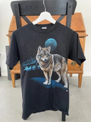Svart t-shirt med vargtryck och måne - Cool svart t-shirt med ett stort tryck av en varg framför en blå måne och skog. T-shirten har rund hals och korta ärmar. Perfekt för dig som gillar djurmotiv och vill sticka ut med en unik design.