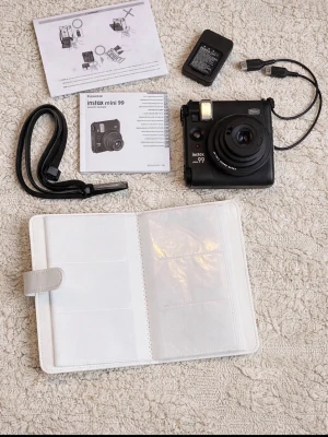 Svart Fujifilm Instax Mini 99 kamera - Säljer en svart Fujifilm Instax Mini 99 direktfilmskamera. Kameran har en kompakt, fyrkantig design i plast och levereras med handledsrem, laddare, USB-kabel, manualer och ett vitt fotoalbum. Perfekt för att ta analoga bilder direkt och spara minnen på papper.
