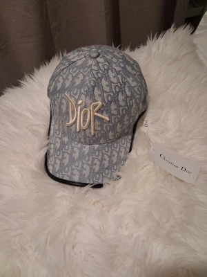 Grå mönstrad keps från Dior - Säljer en grå keps från Christian Dior med ikoniskt monogrammönster och broderad Dior-logga framtill. Kepsen har svarta detaljer längs kanten och är tillverkad i bomullsmix. Perfekt accessoar för en trendig och exklusiv look.