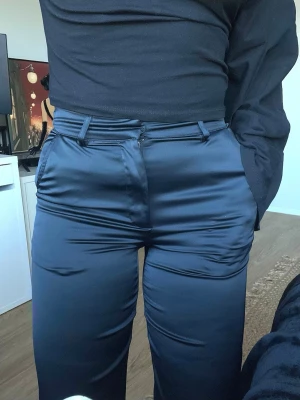 Marinblå glansiga kostymbyxor Bikbok - nyskick, strl 34 - Marinblå glansiga kostymbyxor med raka ben. Hällor för skärp samt fuskfickor bak. Polyester och elastan. Använda knappt 5 gånger - nyskick!