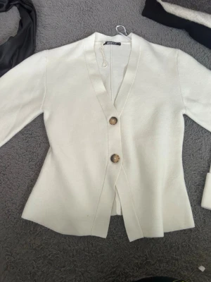 Vit kofta med knappar från Gina Tricot - Stilren vit kofta från Gina Tricot med två stora bruna knappar framtill. Koftan har v-ringning, lång ärm och lätt figursydd passform. Tillverkad i mjukt stickat material som ger en clean och modern look.