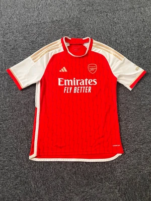 Arsenal matchtröja röd Adidas - Snygg röd Arsenal matchtröja från Adidas med vita och guldiga detaljer på ärmarna. Tröjan har klubbmärke och Adidas-logga på bröstet samt Emirates Fly Better-tryck framtill. Tillverkad i lätt och ventilerande funktionsmaterial, perfekt för fotboll eller träning. Helt Äkta!