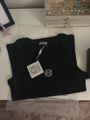 Svart ärmlös linnetopp från Loewe - Snygg svart ärmlös linnetopp från Loewe med rund halsringning och vit broderad logga på bröstet. Perfekt att styla med jeans eller kjol för en clean och trendig look. Materialet känns mjukt och följsamt. Finns i vit och svart, 3 veckor leverans. Alla storlekar 