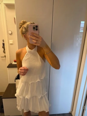 Vit volangklänning med halterneck - Supersöt vit kortklänning med halterneck och flera lager volanger. Klänningen är ärmlös och har öppen rygg med knyt i nacken. Perfekt för sommarens alla tillfällen och ger en fräsch och trendig look.