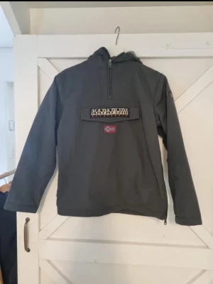 Grå  vindjacka från Napapijri - Stilren grå vindjacka från Napapijri med huva och halv dragkedja framtill. Stor ficka på bröstet med Napapijri-logga och norsk flagga. Jackan har en sportig look och är tillverkad i slitstarkt material, perfekt för blåsiga dagar.
