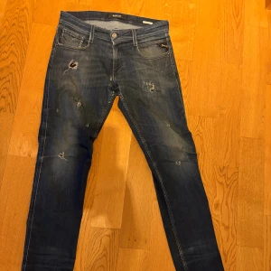 Blå slitna jeans från Replay - Säljer ett par blå jeans från Replay med skinny fit och slitningar för en trendig, avslappnad look. Jeansen har klassisk femficksmodell, dragkedjegylf och detaljerade slitningar på flera ställen. Perfekta för dig som gillar streetstyle.