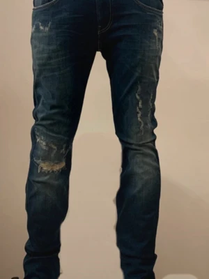 Replay Anbass blå jeans med slitningar  - Säljer ett par Replay Anbass jeans i blå tvätt med slitningar och distressed detaljer på benen. Klassisk femficksmodell med raka ben och snygga kontrastsömmar. Märkespatch bak i midjan och cool faded effekt framtill. Perfekta för dig som gillar en avslappnad men trendig look. Endast använd fåtal gånger utan några som helst defekter. 