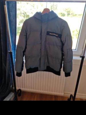 Grå och svart pufferjacka Calvin Klein - Snygg pufferjacka från Calvin Klein i grått och svart med huva och dragkedja. Jackan har en logotyp på bröstet och breda muddar vid ärmslut och nederkant. Perfekt för kalla vinterdagar och riktigt skön att bära.