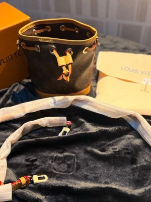 L-V Monogram Bucket Bag - Säljer en snygg bucket bag i klassiskt brunt monogrammönster med detaljer i gult och rött. Väskan har guldfärgade metalldetaljer, dragsko och avtagbar axelrem. Kommer med dustbag och box samt en liten sjal som ni kan se på bild 2. Perfekt för dig som gillar lyxiga accessoarer.