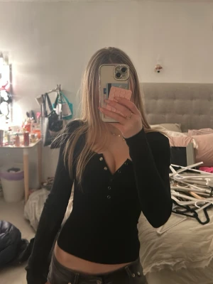 Svart långärmad topp med knappar - Snygg svart långärmad topp med v-ringning och knappar framtill. Figurnära passform som framhäver siluetten. Perfekt att styla med jeans eller kjol för en trendig look.