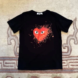 Svart Comme des Garçons PLAY t-shirt - Säljer en svart t-shirt från Comme des Garçons PLAY med det ikoniska röda hjärtat med ögon på bröstet. T-shirten har korta ärmar och en klassisk passform. Perfekt för dig som vill sticka ut lite extra.
