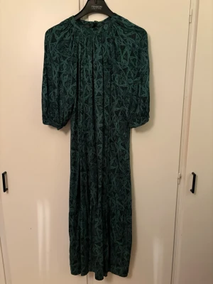 Grön mönstrad långklänning - Snygg långklänning i mörkgrönt mönster.. Klänningen har puffiga halvlånga ärmar och rund halsringning med knytning i nacken. Är snabb att klippa bort lappar. Men den är endast använd 2 ggr. Så absolut som ny! Kostade över 1000kr nypris.