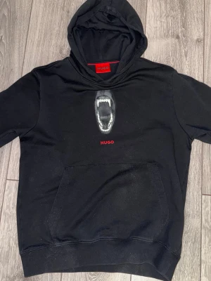 Svart hoodie från Hugo med tryck - Svart hoodie från Hugo med ett coolt tryck av ett öppet gap med tänder på bröstet och röd Hugo-logga under. Klassisk känguruficka framtill och huva. Perfekt för dig som gillar streetwear och vill sticka ut lite extra