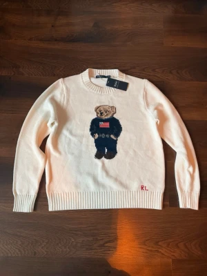 Vit stickad tröja Polo Ralph Lauren - Vit stickad tröja från Polo Ralph Lauren med ikonisk björn på bröstet. Tröjan har rund halsringning, ribbade muddar och RL-broderi nedtill. Björnen bär en blå outfit med amerikansk flagga. Perfekt för dig som gillar klassisk preppy stil.
