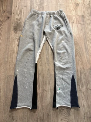 Gallery Dept. - Säljer ett par grå mjukisbyxor från Gallery Dept. med svart logga på vänster sida. Byxorna har elastisk midja, svarta detaljer vid bensluten och färgstänk i vitt, gult och grönt för en unik look. Perfekta för en avslappnad och trendig stil. Storlek L passar bättre på S/M