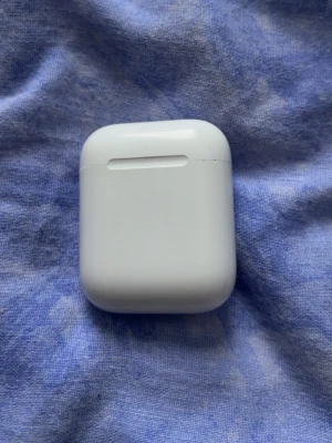 Apple AirPods med laddningsetui - Säljer ett par vita Apple AirPods med det klassiska laddningsetuiet. Etuiet är kompakt, har rundade hörn och är gjort i blank plast. Perfekt för trådlös musik och samtal, smidig att ta med överallt.