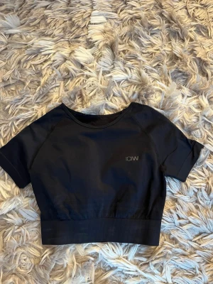 Svart croppad topp från ICANIWILL - Snygg svart croppad topp från ICANIWILL med korta ärmar och diskret logga på bröstet. Tillverkad i ett stretchigt syntetmaterial som passar perfekt till träning. Enkel och stilren design med ribbad kant nedtill.