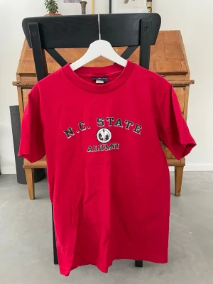 Röd N.C. State Alumni t-shirt - Snygg röd t-shirt med trycket 'N.C. STATE ALUMNI' och ett emblem på bröstet. Klassisk rund hals och korta ärmar. Perfekt för dig som vill ha en college-inspirerad look. Tillverkad i mjuk bomull för skön känsla.