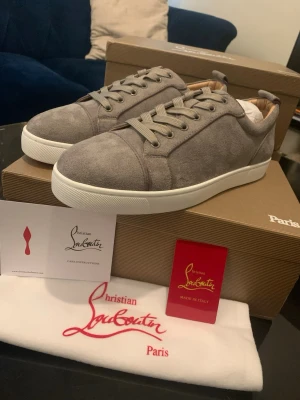 Gråa sneakers från Christian Louboutin - Stilrena gråa sneakers i mocka från Christian Louboutin med klassisk röd sula och vita kanter. Skorna har lågt skaft, matchande grå skosnören och beige innerfoder. Perfekta för dig som vill ha en lyxig och trendig look.