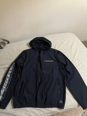 Mörklblå Lacoste vindjacka med huva - Snygg blå vindjacka från Lacoste med huva och dragkedja framtill. Jackan har två fickor med dragkedja och en stor vit Lacoste-logga på vänster ärm samt en liten logga vid höften. Helt ny använd 1 gång , nypris 3000 