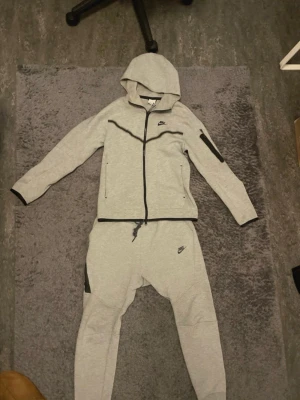 Grå Nike tech fleece tracksuit från Nike - Gråa mjukisbyxor från Nike i mjukt sweatmaterial med svart Nike-logga på benet. Byxorna har dragsko i midjan och svarta detaljer vid fickorna. Matchande hoodie med dragkedja och huva samt svarta dragkedjor och logga. Perfekt för chill eller träning.