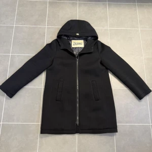 Svart kappa med huva från Herno - Stilren svart kappa från Herno med huva och dragkedja framtill. Kappan har raka ärmar, två fickor och en minimalistisk design. Perfekt för dig som gillar klassisk och enkel stil. Materialet ser ut att vara mjukt och något vadderat.
