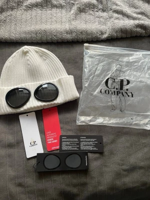 Vit Goggle-mössa från C.P. Company - Vit ribbstickad mössa från C.P. Company med ikoniska svarta goggle-linser på uppviket. Mössan är tillverkad i mjukt material och har en modern, ungdomlig design som sticker ut. Perfekt för dig som vill ha en unik accessoar med streetwear-vibe.