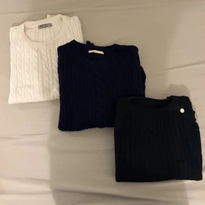 Stickade tröjor i vitt, marinblått och svart - Tre stickade tröjor i klassisk kabelstickad design. En vit, en marinblå och en svart tröja, alla med rund halsringning och ribbade muddar. Perfekta för lager på lager och en stilren look.