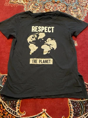 Svart t-shirt med tryck RESPECT - Cool svart t-shirt med beige tryck på bröstet som visar världskartan och texten 'RESPECT THE PLANET'. Klassisk rund hals och korta ärmar. Perfekt för dig som vill göra ett statement med din stil.