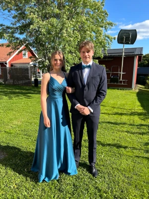 Blå långklänning / balklänning  - Säljer en elegant blå långklänning med v-ringning och smala axelband. Klänningen har öppen rygg med snygg snörningsdetalj och är tillverkad i ett glansigt, mjukt material som faller fint. Perfekt för bal eller student. Det finns diskreta fläckar på den som självklart skickas vid förfrågan. 