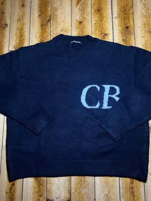 Svart stickad tröja Cole Buxton - Svart stickad tröja från Cole Buxton med stora vita bokstäver ”CB” på framsidan. Tröjan har rund halsringning, ribbade muddar vid ärmslut och nederkant samt en avslappnad passform. Perfekt för dig som gillar streetwear och enkel design men även gillar exklusiva kläder