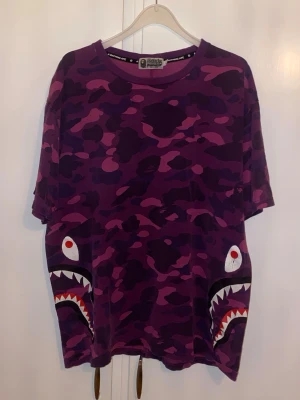 Lila camo t-shirt från BAPE - Säljer en lila camo t-shirt från A Bathing Ape med ikoniska haj-motiv på sidorna. Köpt på Bape butik i Tokyo - Japan. 