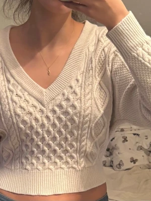 Beige kabelstickad croppad tröja - Trendig beige kabelstickad tröja med croppad passform och djup V-ringning. Tröjan har långa ärmar, bred ribbad mudd och en snygg knytning i ryggen. Materialet är mjukt och tjockt, perfekt för lager på lager. Snygga stickade detaljer över hela tröjan.