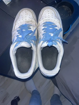 Nike Air Force 1 vita/blå sneakers - Nike Air Force 1 sneakers i vitt läder med blå detaljer och blå skosnören. Klassisk låg modell med perforerad tåbox och tydlig Swoosh-logga på sidan. Yttersulan är vit med blå kant. Perfekta för dig som gillar retrostil och streetwear.