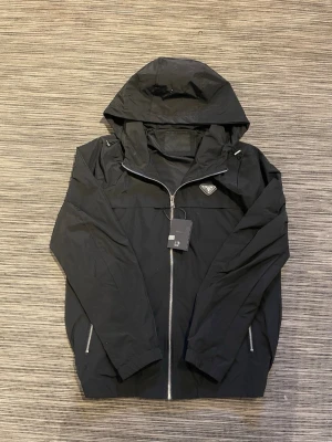 Svart vindjacka från Prada med huva - Säljer en snygg vindjacka/Windbreaker från Prada! Jackan är i mycket bra skick och har inga defekter. Luvan går att spänna. Om ni har några frågor så är de bara att höra av er!