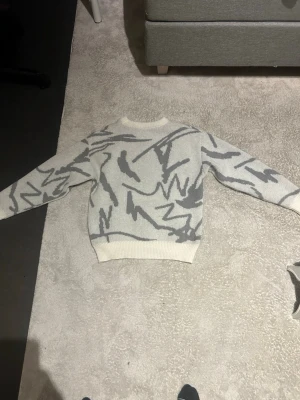 Stickad tröja  - Säljer en stickad tröja i vit och grå färg med coolt abstrakt mönster. Tröjan har rund hals och långa ärmar med ribbade muddar vid hals, ärmslut och nederkant. Perfekt för dig som gillar unik stil och mjuka material.