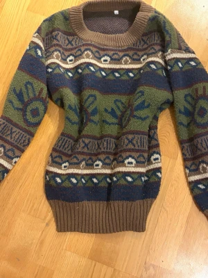 Retro mönstrad stickad tröja - Stickad tröja med retroinspirerat mönster i grönt, blått, brunt och beige. Tröjan har rund halsringning och ribbstickade muddar vid ärmslut och nederkant. Perfekt för dig som gillar vintagevibbar och unika plagg.
