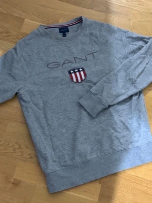 Grå sweatshirt från GANT - Snygg grå sweatshirt från GANT med broderad logga och ett emblem med stjärnor och ränder på bröstet. Tröjan har rund halsringning och långa ärmar. Tillverkad i mjuk bomull för skön känsla. Jätte bra skick. Storlek: S
