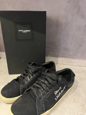 Svarta Saint Laurent sneakers - Svarta sneakers från Saint Laurent med vit logga på sidan. Skorna har låga skaft, svarta skosnören och vit sula. Ovandelen är i canvas och detaljer i läder. Klassisk och stilren design med en edgy touch.