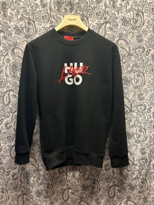 Svart sweatshirt från Hugo - Stilren svart sweatshirt från Hugo med vit och röd logga framtill. Tröjan har rund halsringning, långa ärmar och ribbade muddar vid ärmslut och nederkant. Perfekt för en trendig och avslappnad look.