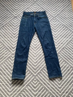 Mörkblå jeans från Lee - Säljer ett par klassiska mörkblå jeans från Lee med fin passform och fem fickor. Jeansen har snygga kontrastsömmar och metallknapp i midjan. Perfekta till vardagslooken och tillverkade i slitstarkt denimtyg. Väldigt schyssta slitningar. Storlek W29 L34 Men skulle säga att Längden passar ner till 32 beroende på hur man vill att dem ska sitta👍